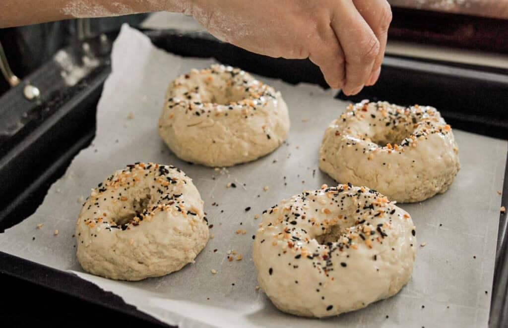 A persons hand sprinkles seasoning onto four uncooked bagels resting on a parchment-lined baking sheet. The bagels are topped with a mix of black and white seeds. Flour dust is visible on the fingers.